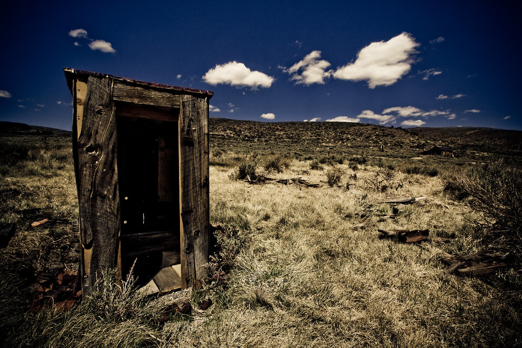 A wooden outhouse sits in the left corner of a desert scene with blue sky and sage brush