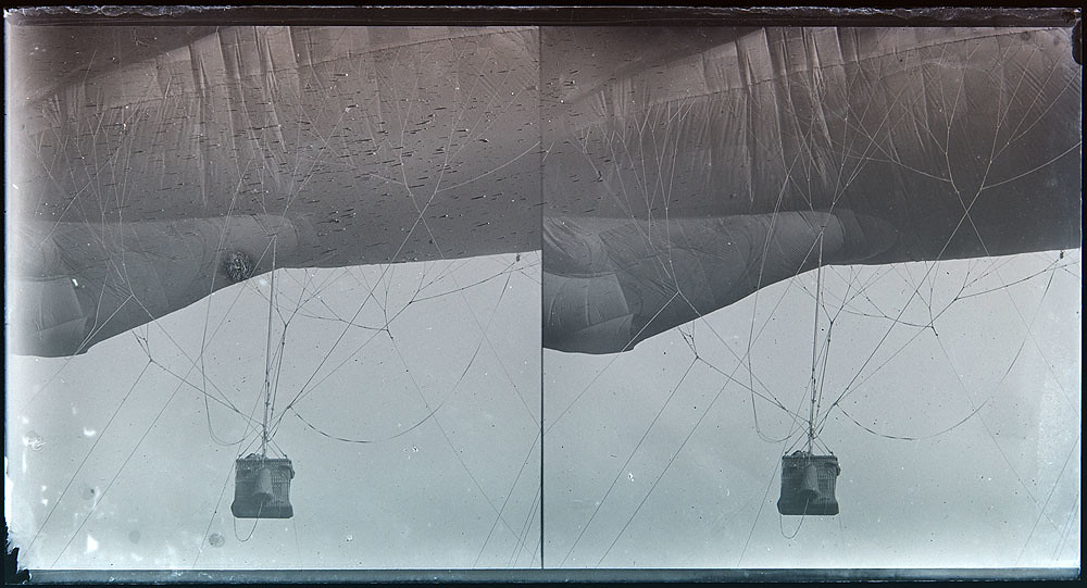 A black and white stereoscopic card of a WWI-era observation balloon - a small basket with a single person visible inside in dangles precariously from a hot air balloon