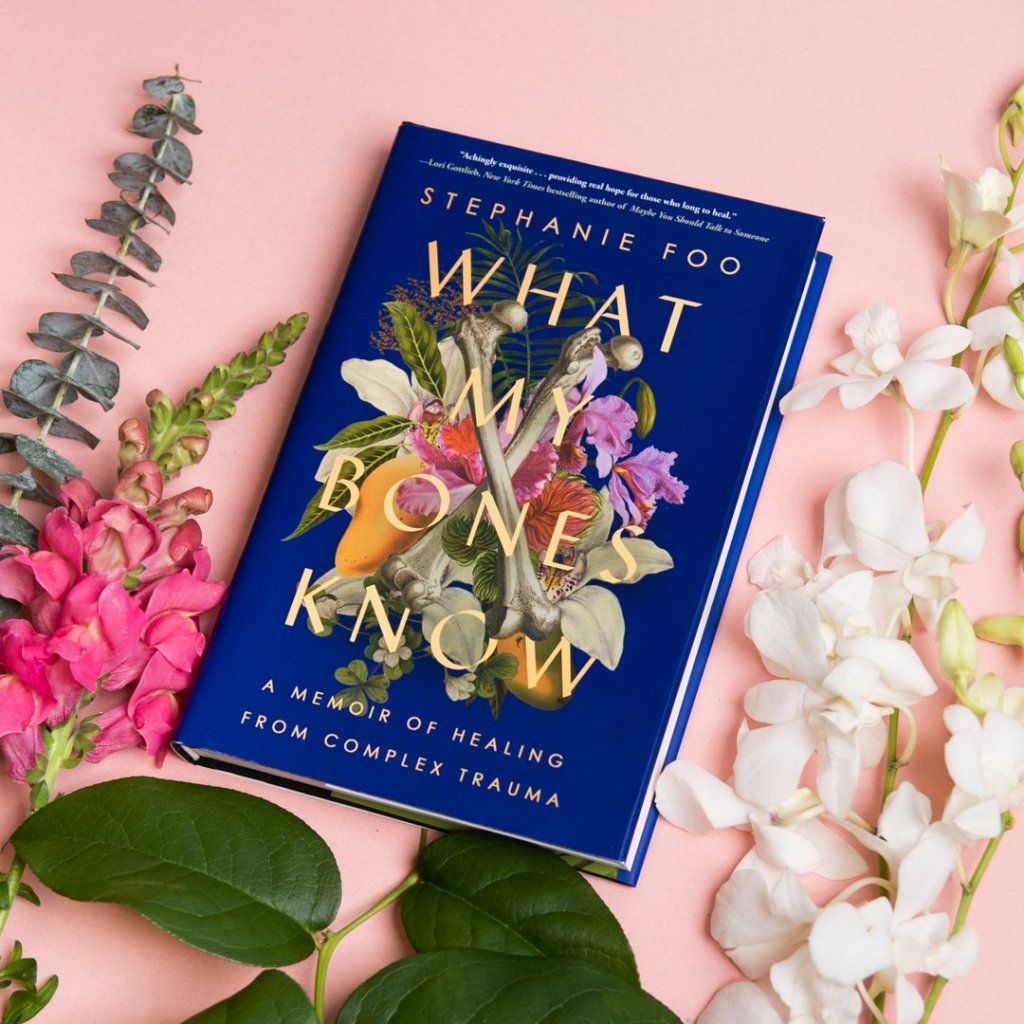 Stephanie Foo's "What My Bones Know: A Memoir of Healing from Complex Trauma" sits on a blush pink background, surrounded by flowers. The book's cover is a deep purple and contains an illustration of still life of flowers, fruit, and what appear to be human femur bones.